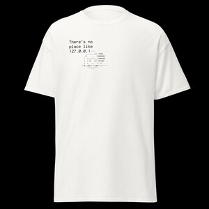 May include: White t-shirt with short sleeves. The shirt features the text "There's no place like 127.0.0.1" in black, along with a stylized graphic. The design is located on the upper left chest area of the shirt.