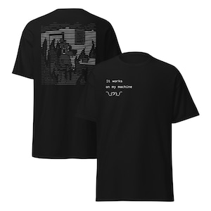 Può includere: T-shirt nere con grafica bianca. Sul davanti si legge "It works on my machine" con uno shrug ASCII. Il retro della maglietta presenta un disegno pixelato di paesaggio urbano in bianco.