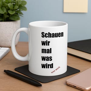 May include: White ceramic mug with the German phrase "Schauen wir mal was wird" printed in black, with "was wird" in red. The mug sits on a black square coaster, with a black pen and a smartphone nearby.