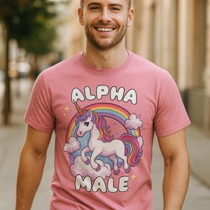 Puede incluir: Camiseta rosa con las palabras "ALPHA MALE" arriba y abajo de un unicornio de dibujos animados con un arcoíris y nubes. El unicornio tiene una melena y cola coloridas. La camiseta es de manga corta.