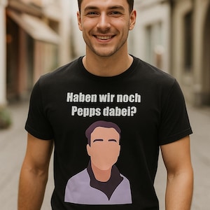 May include: Black crew neck t-shirt with the German text "Haben wir noch Pepps dabei?" in white. The shirt features a cartoon illustration of a person in a purple jacket. The person in the image is smiling.