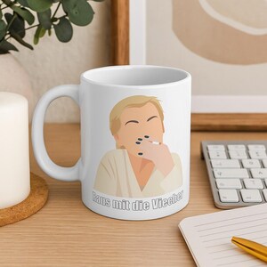 Puede incluir: Taza de cerámica blanca con una ilustración de dibujos animados de una persona con cabello rubio, sosteniendo un cigarrillo. La taza tiene el texto "Raus mit die Viecher" en negro.