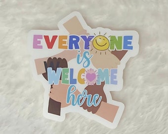 Può includere: Un adesivo colorato con il testo "Everyone is welcome here" e una grafica di mani che si tengono in cerchio. Le mani sono di colori diversi, a rappresentare la diversità. Un sole sorridente è in cima al cerchio.
