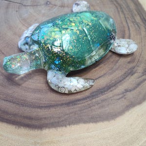 Peut inclure: Une figurine de tortue en résine verte avec des paillettes incrustées dans la résine. La tortue est posée sur une surface en bois.