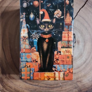 Peut inclure: Un chat noir portant un bonnet de Père Noël rouge est assis sur une pile de cadeaux emballés colorés. L'arrière-plan est bleu foncé avec des étoiles et des ornements.