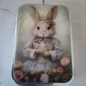 Peut inclure: Boîte rectangulaire avec une illustration vintage d'un lapin portant une couronne florale et une robe, entouré de roses. Le lapin a une expression sereine. La boîte a une bordure argentée et est posée sur un fond en bois clair.