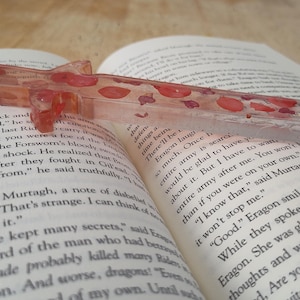 May include: A clear, pink bookmark with red and pink flowers embedded inside. The bookmark is laying on top of an open book.