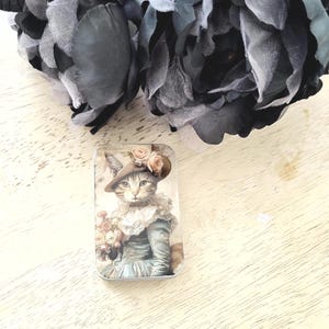 May include: A rectangular metal tin featuring a vintage illustration of a cat wearing a hat adorned with roses and a blue dress, holding a bouquet. The tin is set against a light wooden surface, with dark gray fabric flowers in the background.