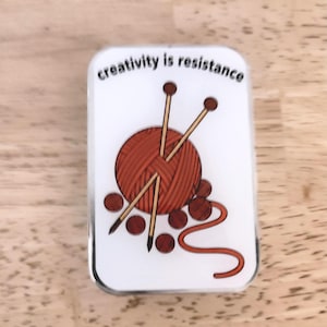 May include: A rectangular white pin with a silver border. The pin features a red yarn ball with two knitting needles, several small red circles, and the text "creativity is resistance" in black.