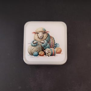 Pode incluir: Base quadrada com uma ilustração de uma ovelha a tricotar com novelos de lã em tons de bege, azul e laranja. A ovelha usa um suéter azul e estampado. Uma ovelha menor com um suéter a condizer também está presente. A base tem um fundo branco.