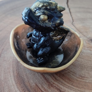 May include: A small black and gold dragon figurine sitting in a gold bowl. The dragon has a detailed face and wings.