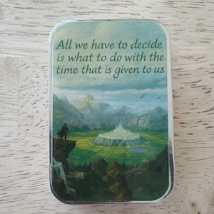 May include: Rectangular tin with a landscape scene featuring mountains, a castle, and a figure on a cliff. The tin has the quote, "All we have to decide is what to do with the time that is given to us."