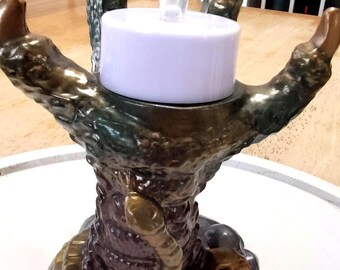 May include: A ceramic candle holder shaped like a hand with a snake and skull at the base. The hand is holding a white candle with a clear flame.