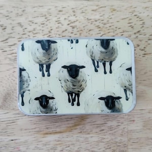 May include: Rectangular tin with a repeating pattern of sheep. The sheep have white wool and black faces and legs. The background is a light beige color with vertical lines. The tin has a silver edge.