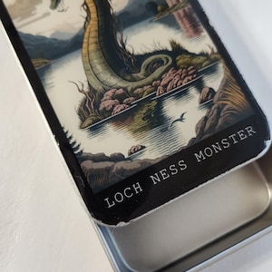 May include: A rectangular tin featuring an illustration of the Loch Ness Monster in a lake with mountains and a castle in the background. The text "LOCH NESS MONSTER" is printed on the bottom edge of the tin.