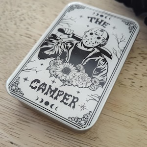 May include: A rectangular, silver-colored metal tin featuring a black and white illustration of a masked figure holding a weapon, with the text "THE CAMPER". Floral and celestial design elements surround the central image.