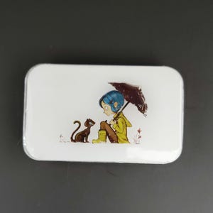 May include: A white rectangular tin featuring a cartoon illustration. The image depicts a girl with blue hair and a yellow raincoat sitting with a black cat under a black umbrella. The artwork has a whimsical, storybook style.