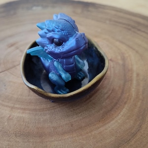 May include: A small blue and purple dragon figurine sitting in a brown ceramic bowl. The dragon has a detailed face and wings.