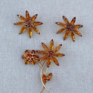 Peut inclure: Un ensemble de bijoux composé d'une broche et de deux boucles d'oreilles assorties. Chaque pièce présente des cristaux ambrés taille marquise disposés en motif floral avec un petit cristal rouge au centre, sur une monture dorée avec tiges.