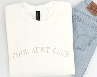 May include: A white crewneck sweatshirt featuring the text COOL AUNT CLUB embroidered in a light beige thread across the chest. The sweatshirt is paired with light blue denim jeans that have a brown leather patch visible on the back pocket.