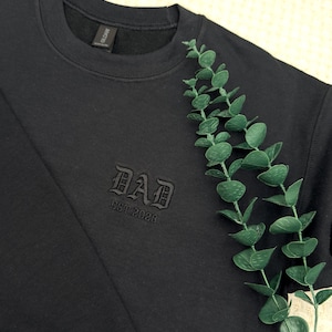 May include: Black crewneck sweatshirt with the word "DAD" embroidered in a gothic font. The text "EST. 2026" is embroidered below. A sprig of green eucalyptus is placed on the right side of the sweatshirt.