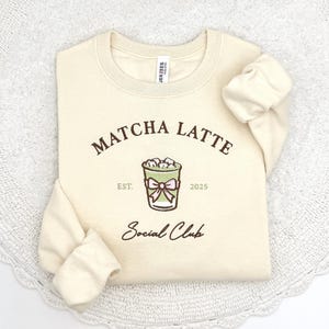 May include: Cream-colored sweatshirt with the words "Matcha Latte" in brown script above a green and pink graphic of a matcha latte. The text "Est. 2025" is below the graphic, and "Social Club" is written in script below.