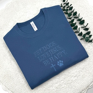 May include: A blue crewneck sweatshirt featuring the embroidered text PET DOGS. LOVE JESUS. BE HAPPY. along with a small embroidered cross and a dog paw print icon. The garment includes a white interior neck label with the brand name Jerzees.