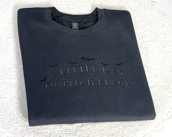 May include: A black crewneck sweatshirt featuring tonal black embroidery on the chest. The text reads A LITTLE RAY OF PITCH BLACK, accompanied by small embroidered bat silhouettes positioned above the lettering in a curved arrangement.