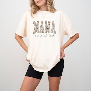 May include: An off-white short-sleeve t-shirt featuring the word MAMA in large block letters filled with a floral pattern in shades of brown, green, and pink. Below the word MAMA, the text optional text is printed in a black cursive font.