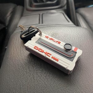 May include: A silver and red key fob with the text "12 VALVE DOHC 3000" is resting on a black surface. The key fob has a black circular design and a key attached to a silver ring.