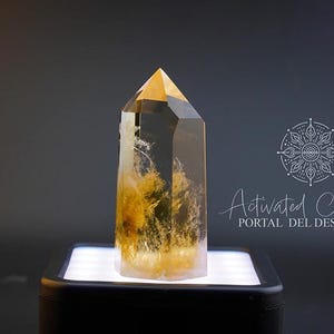 Puede incluir: Una punta de cristal de cuarzo transparente con un tono amarillo dorado. El cristal está sobre una plataforma blanca con una base negra. El texto "Activated Crystals Portal del Despertar" es visible en el fondo.