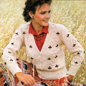 May include: A cream-colored knitted cardigan with a V-neck and brown floral embroidery. The cardigan is worn over a red button-down shirt and a brown beaded necklace.