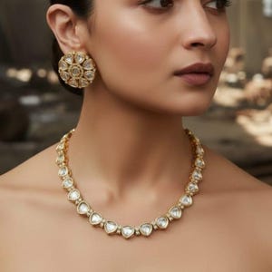 May include: A gold-toned necklace and matching earrings set. The necklace features heart-shaped clear stones set in gold. The earrings are round with a similar design. The jewelry is worn on a person with dark hair and a neutral-toned top.