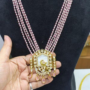 May include: A gold-toned necklace and earring set featuring a pendant with a white inlay and a gold bird design. The necklace has multiple strands of pink beads, and the earrings match the pendant design. The set is displayed on a black mannequin.