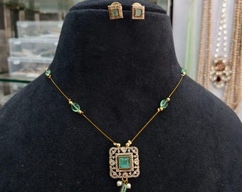 May include: A gold-toned necklace and matching earrings set. The necklace features a square pendant with a central green gemstone, surrounded by smaller stones. The earrings are square-shaped with a green gemstone. The jewelry is displayed on a black mannequin.