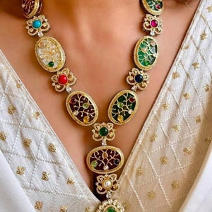May include: A gold-toned statement necklace featuring oval and round gemstone pendants in various colors, including green, red, and blue. The necklace has a floral design and a large green pendant at the bottom. The necklace is worn over a white garment.