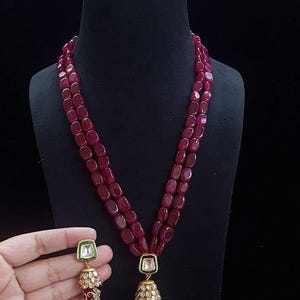Op de afbeelding: Een ketting met drie strengen en bijpassende oorbellen. De ketting heeft ovale, dieprode kralen. De hanger en oorbellen hebben een vierkante, witte steen met gouden en parelaccenten. De oorbellen worden door een hand vastgehouden.