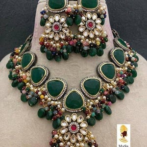 May include: A green gemstone necklace and earring set. The jewelry features large, teardrop-shaped green stones, floral accents, and dangling beads in various colors. The earrings and necklace are displayed on a mannequin.