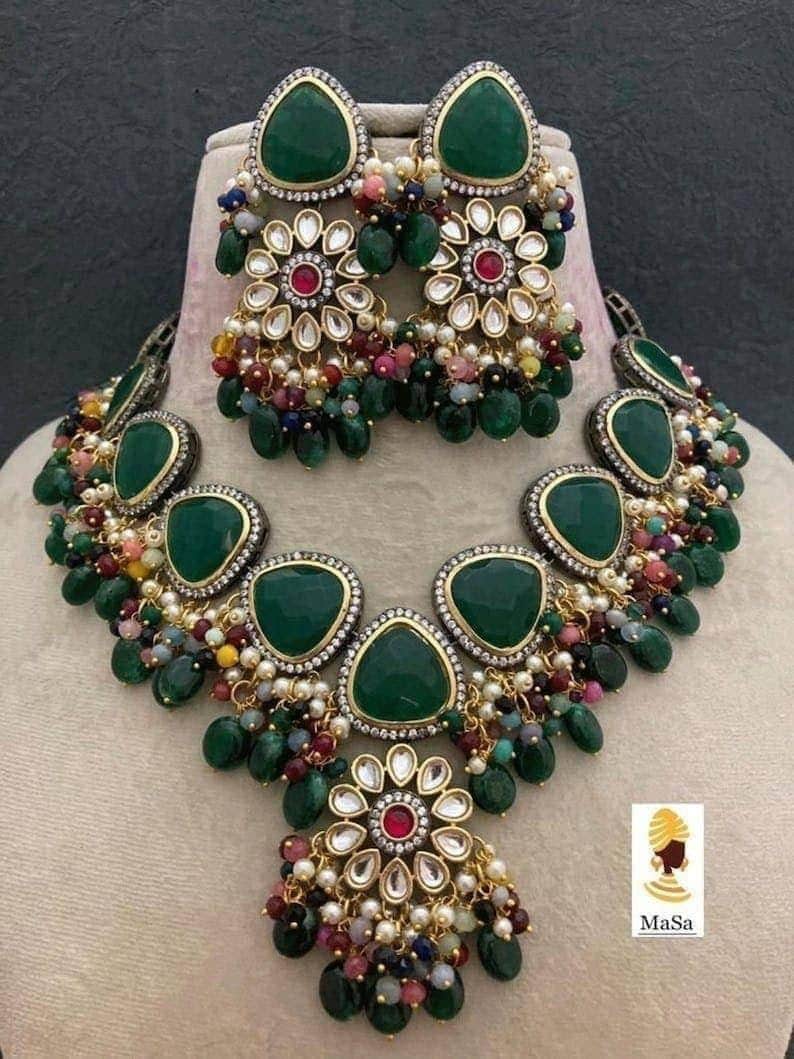 May include: A green gemstone necklace and earring set. The jewelry features large, teardrop-shaped green stones, floral accents, and dangling beads in various colors. The earrings and necklace are displayed on a mannequin.