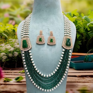 May include: A necklace and earring set displayed on a mannequin. The necklace features a large, rectangular green stone pendant with a gold-colored frame, surrounded by clear stones. The necklace has multiple strands of green beads and white pearls. The earrings match the necklace.