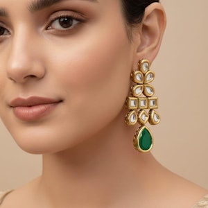 Puede incluir: Pendientes colgantes dorados con un diseño geométrico. Los pendientes presentan detalles claros, cuadrados y con forma de flor, y una piedra verde esmeralda en forma de lágrima en la parte inferior. Los pendientes se llevan en la oreja.