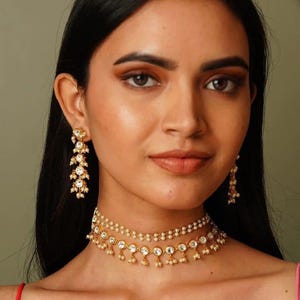 May include: A woman wearing a gold-toned jewelry set. The set includes a layered choker necklace and dangling earrings, both adorned with clear gemstones and small pearls. The jewelry is set against a neutral background.