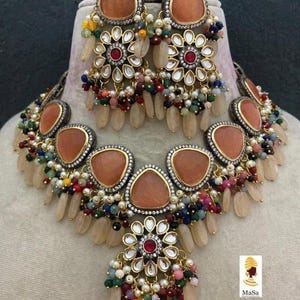 May include: A necklace and earring set featuring peach-colored gemstones and intricate floral designs. The jewelry includes a necklace with multiple peach-colored stones and matching earrings. The set is adorned with colorful beads and white pearls.