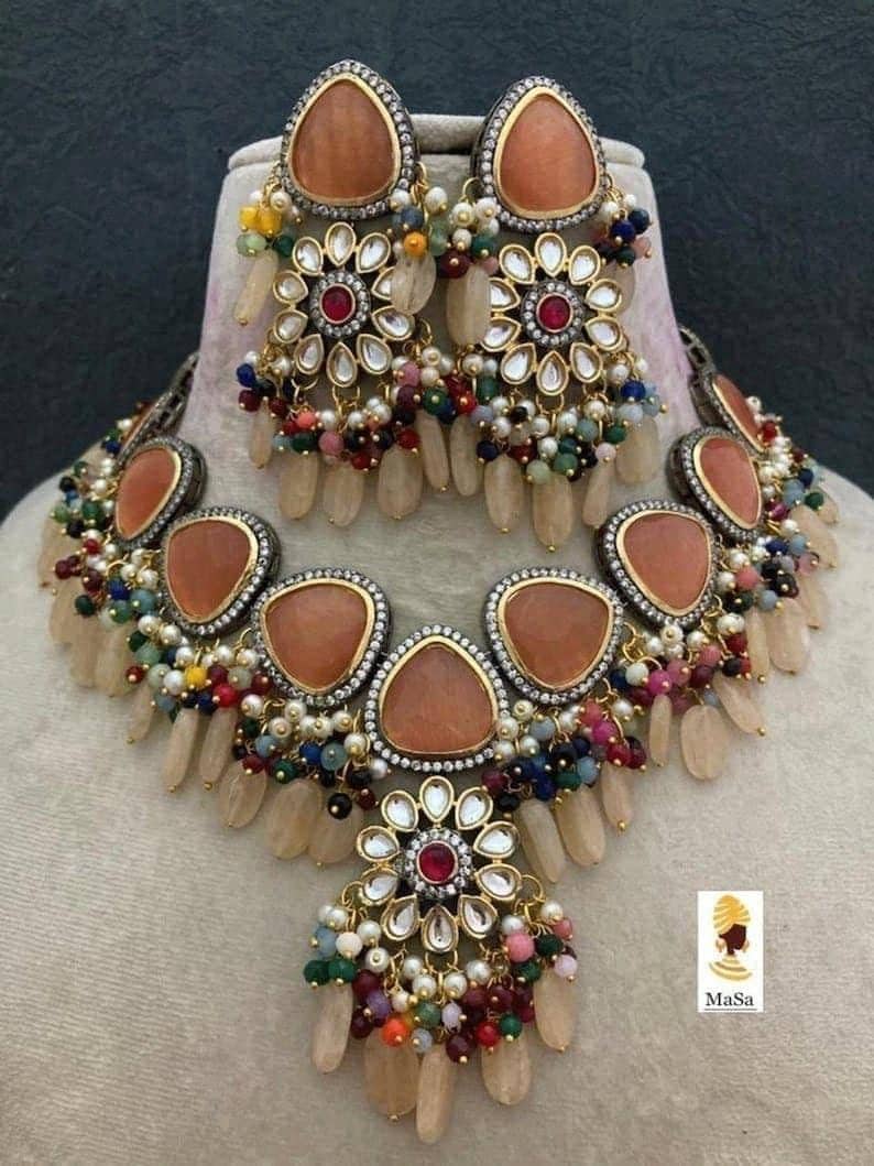 May include: A necklace and earring set featuring peach-colored gemstones and intricate floral designs. The jewelry includes a necklace with multiple peach-colored stones and matching earrings. The set is adorned with colorful beads and white pearls.