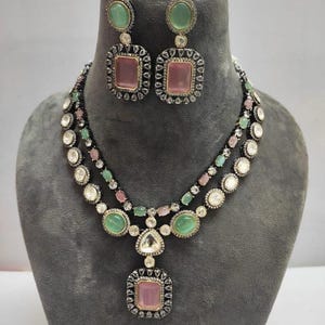Peut inclure: Ensemble de collier et boucles d'oreilles avec des pierres précieuses roses et vertes. Les boucles d'oreilles et le pendentif sont de forme carrée, bordés de petites pierres claires. Le collier est présenté sur un mannequin gris.