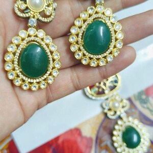 May include: Gold-toned earrings with a floral design. Each earring features a large, oval, emerald-green stone surrounded by small, clear stones. A pearl sits at the top of each earring. The earrings are held up by a hand.