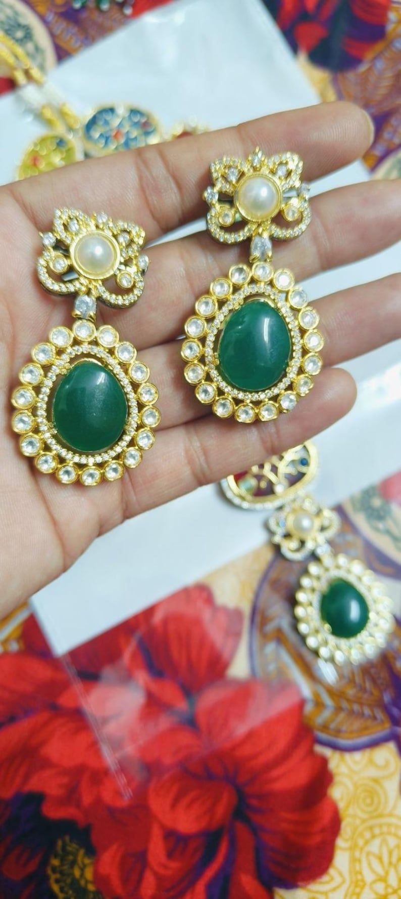 May include: Gold-toned earrings with a floral design. Each earring features a large, oval, emerald-green stone surrounded by small, clear stones. A pearl sits at the top of each earring. The earrings are held up by a hand.