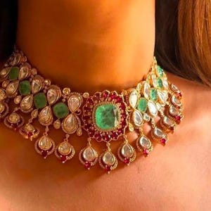 May include: A gold-coloured statement necklace featuring emerald and clear gemstones, with red accents. The necklace has a central emerald stone and teardrop-shaped dangles. The necklace is designed to be worn around the neck.