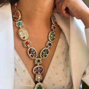 May include: A gold-toned necklace featuring oval-shaped pendants with colorful gemstones and intricate designs. The necklace has a large green teardrop-shaped pendant at the bottom and is worn over a white top.