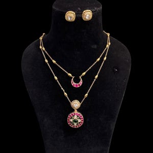 Peut inclure: Un ensemble de bijoux dorés comprenant un collier et des boucles d'oreilles. Le collier comporte deux chaînes, l'une avec un pendentif en forme de croissant et l'autre avec un pendentif circulaire avec des accents rouges et verts. Les boucles d'oreilles sont carrées avec une pierre transparente.